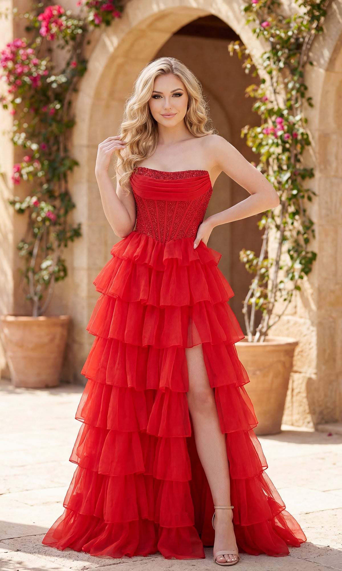 A woman wearing the Long Ruffled Prom Dress: PromGirl Exclusive 250204 in red, with a beaded corset bodice, tiered skirt, and thigh-high slit, stands outdoors near stone arches and potted flowering plants.